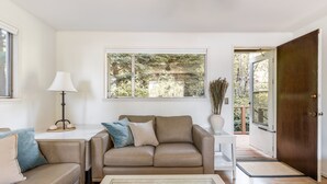 Un salon avec un canapé beige, une table basse en verre et une grande fenêtre donnant sur des arbres.
