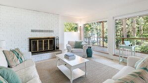 Un salon moderne avec une cheminée en briques blanches, des canapés blancs, une table basse et de grandes fenêtres donnant sur la verdure.
