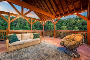 A wooden deck with a wicker sofa and a cushioned chair, surrounded by a wooden railing and string lights.