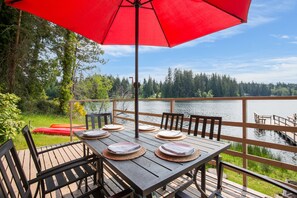 Une terrasse en bois avec une table dressée pour quatre personnes, donnant sur un lac et agrémentée d’un parasol rouge.