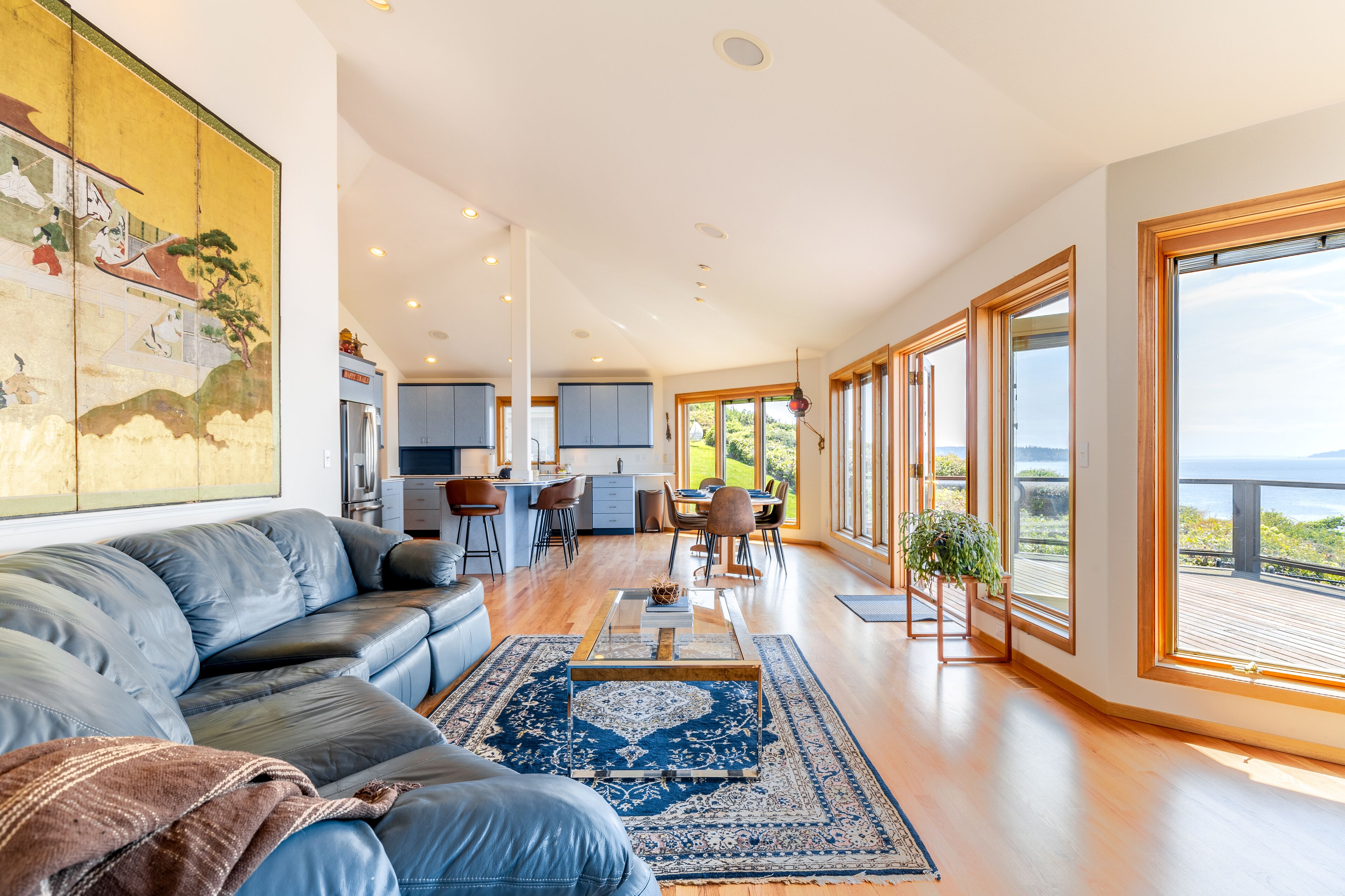 A spacious living area with a leather sofa, a coffee table, and a large rug. There is a kitchen in the background with modern appliances and a dining area with chairs.