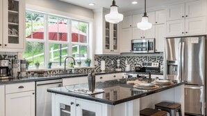 Una cocina moderna con armarios blancos, encimera negra, electrodomésticos de acero inoxidable y un amplio ventanal con vistas a zonas verdes.