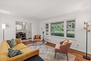 Ein Wohnzimmer mit einem gelben Sofa, zwei rosafarbenen Sesseln, einem Glastisch und einer Stehlampe. Große Fenster bieten Blick auf Grünflächen.