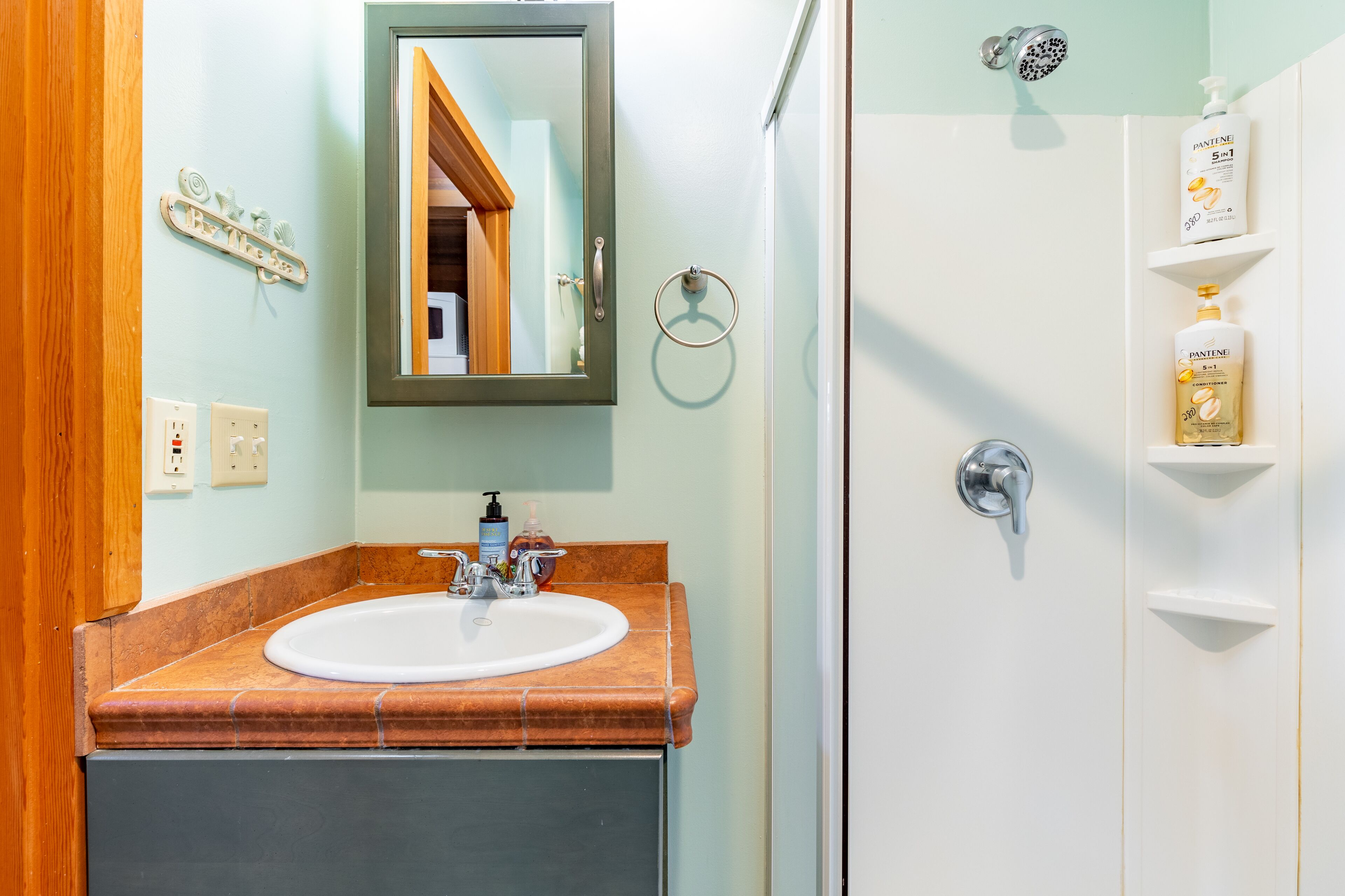 A bathroom with a sink, mirror, towel ring, shower, and toiletries on a shelf.