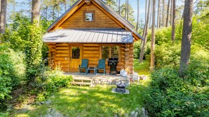En timmerstuga med två stolar på verandan, en grill och en eldstad i sten.