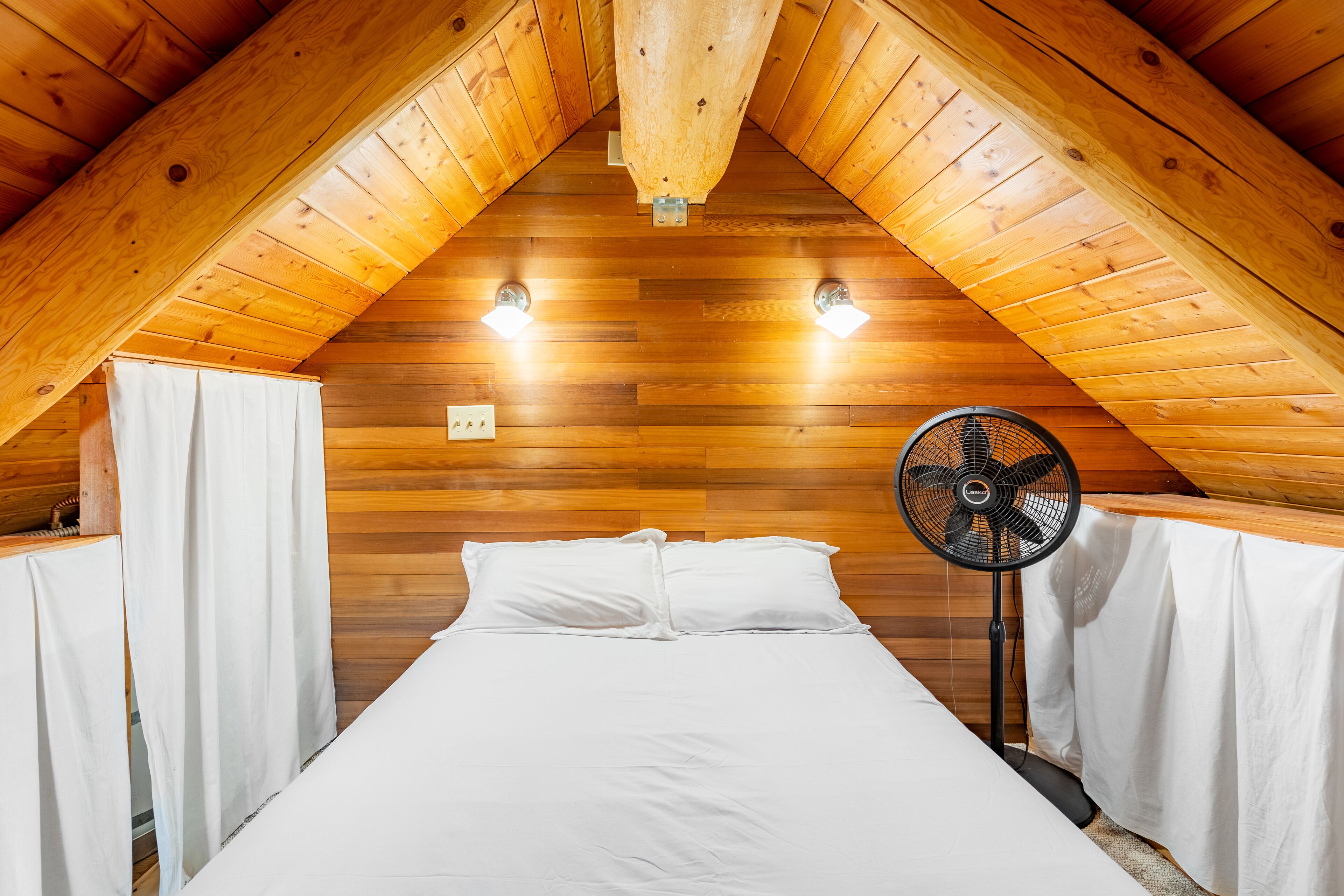 Quarto com painéis de madeira, uma cama, duas luzes fixadas na parede e um ventilador.