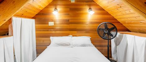Quarto com painéis de madeira, uma cama, duas luzes fixadas na parede e um ventilador.