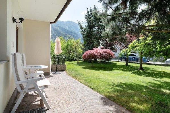 Une terrasse avec des chaises blanches et une table, un petit jardin avec de la pelouse et des arbres, et une vue sur les montagnes en arrière-plan.