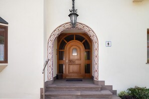 Une porte en bois surmontée d’un arc décoratif, une lampe en métal au-dessus, et une petite marche menant à l’entrée.