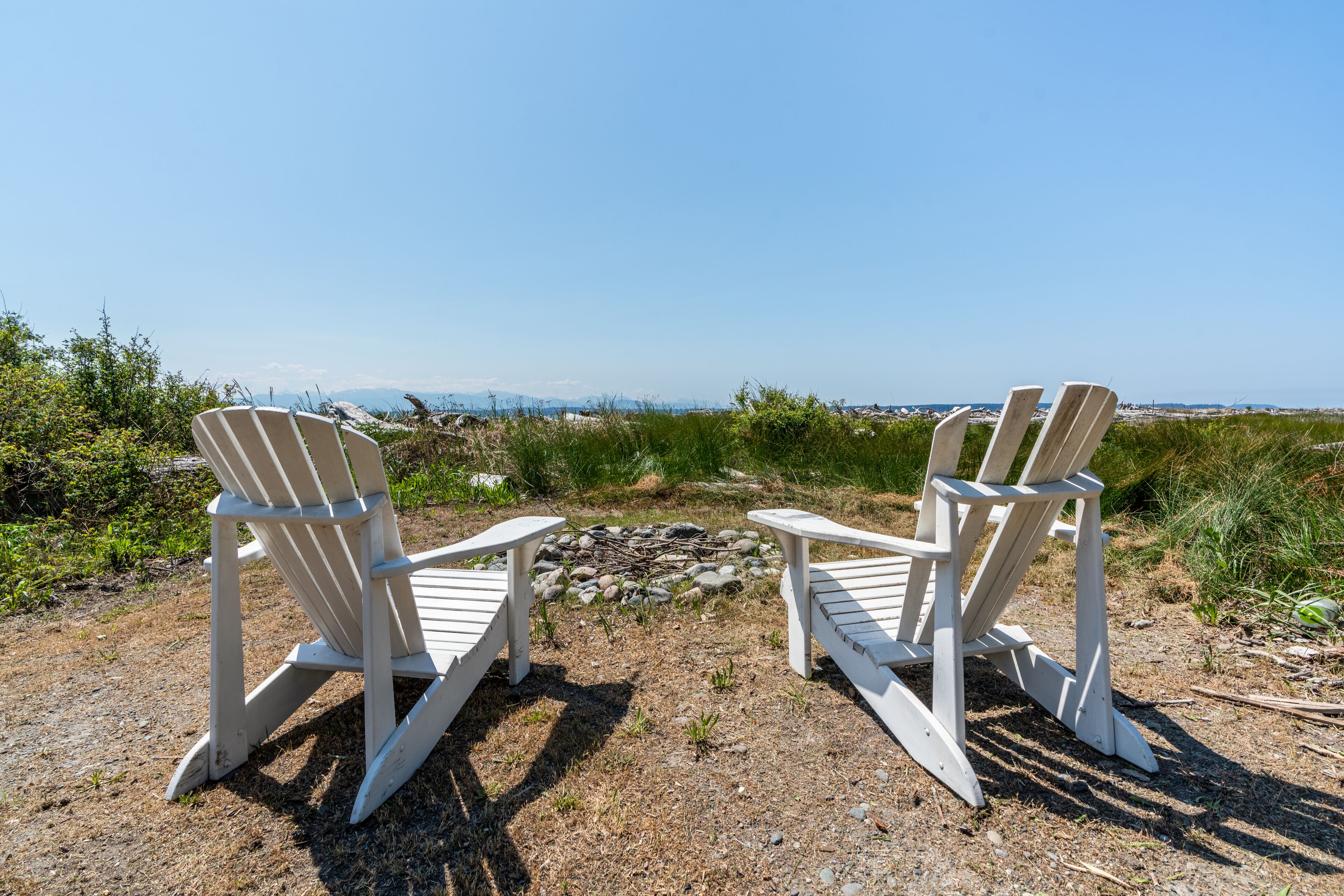 Twee Adirondack-stoelen gericht op een rotsachtig gebied met uitzicht op de oceaan en verre bergen.