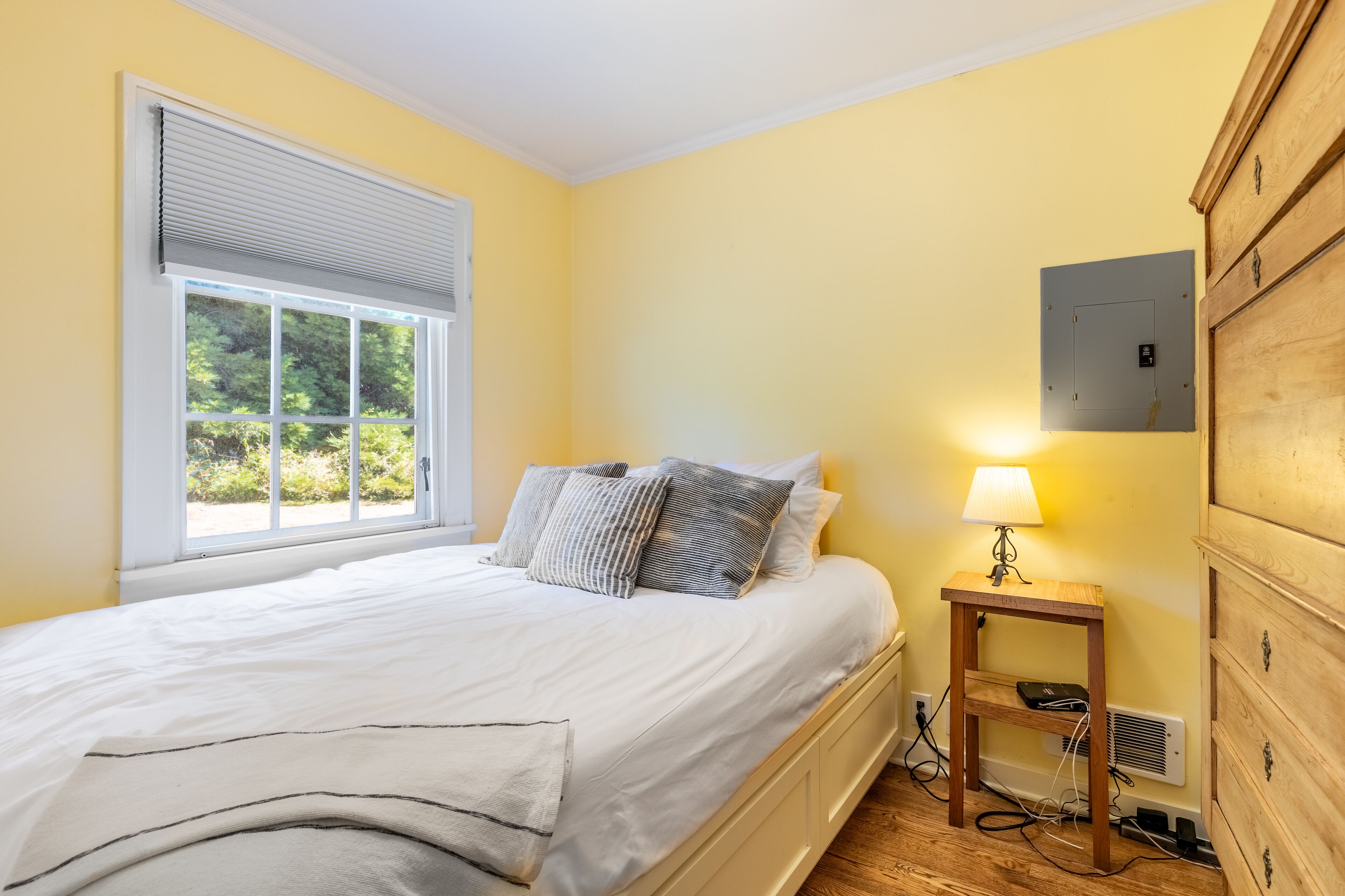 A bedroom with a bed, a bedside table with a lamp, a wooden dresser, and a window with blinds.