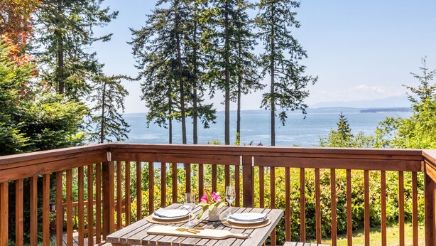 Una terraza de madera con una mesa dispuesta para dos, con vista a un cuerpo de agua y altos árboles.