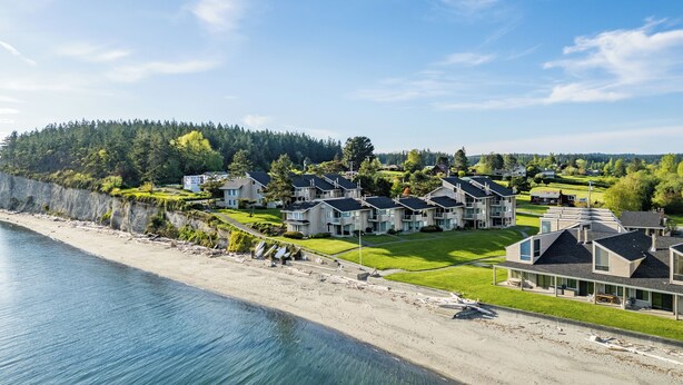 Zona residencial costera con casas modernas, una playa y una colina boscosa al fondo.