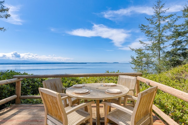 A wooden deck with a round table and chairs, overlooking a body of water and distant mountains.