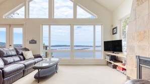 Ein geräumiges Wohnzimmer mit großem Fenster mit Blick auf ein Gewässer, einer Ledersofa, einem Couchtisch und einem Flachbildfernseher.