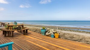 A wooden deck with picnic tables and potted plants overlooking a beach. - West Beach Wonderland by AvantStay | Waterfront (Oak Harbor)