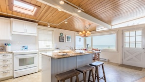 A kitchen with white cabinets, a central island with stools, a large window with a view, and a wooden ceiling. - West Beach Wonderland by AvantStay | Waterfront (Oak Harbor)