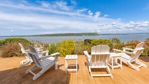 Um deck com quatro cadeiras Adirondack brancas e uma pequena mesa, com vista para a água e terras distantes.