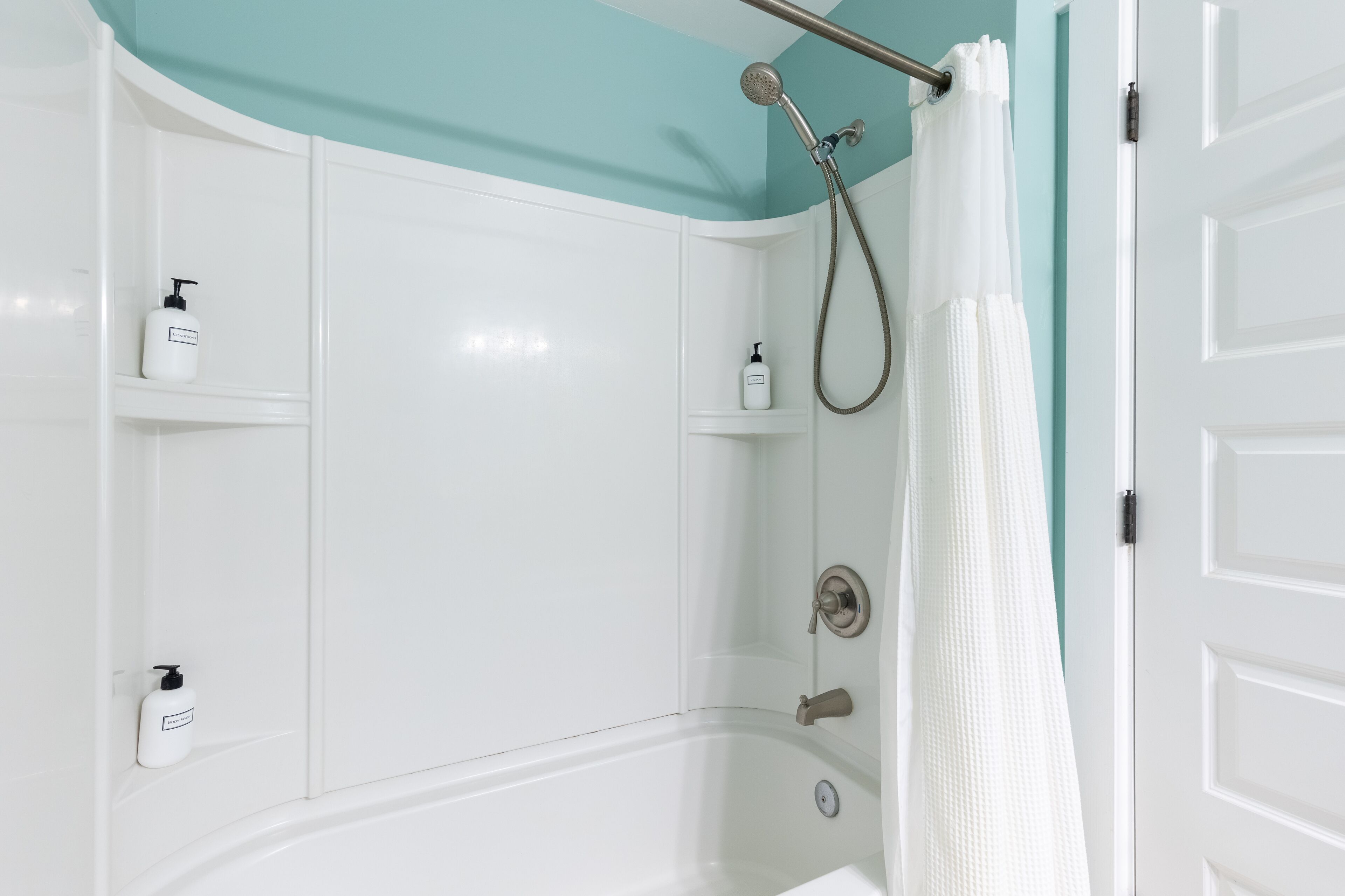 Ein Badezimmer mit einer weißen Badewanne, einer Dusche inklusive Handbrause und zwei weißen Regalen mit Flaschen.