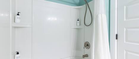 Ein Badezimmer mit einer weißen Badewanne, einer Dusche inklusive Handbrause und zwei weißen Regalen mit Flaschen.