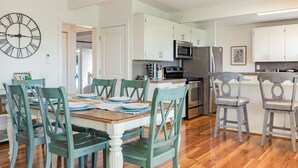 A modern kitchen with white cabinets, a large clock, and a dining area with teal chairs and a wooden table. - Sunny Side Island Retreat by AvantStay | Backyard (Freeland)