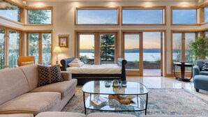 A spacious living room with a large bed, a sofa, and a coffee table. The room has large windows offering a view of a lake and mountains.