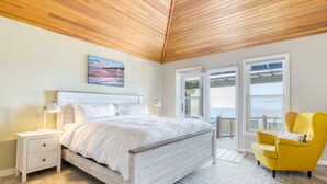 Une chambre avec un plafond en bois, un lit blanc, une table de chevet, un fauteuil jaune et une vue sur l’océan grâce à de grandes fenêtres.