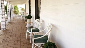Un área de estar al aire libre cubierta con sillas de mimbre blanco y cojines verdes, suelo de ladrillo y pared blanca.
