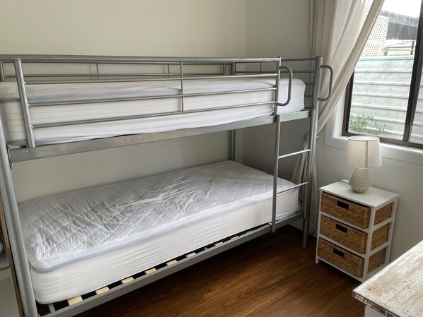 A bunk bed with a white mattress and metal frame, a bedside table with a lamp, and a window with a view of buildings. - Holiday Unit 2 in Port Victoria
(Port Victoria)