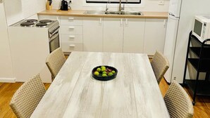 Cocina moderna con armarios blancos, campana extractora de acero inoxidable, horno empotrado, lavamanos y microondas. Hay una mesa de comedor con sillas de mimbre y un bol de fruta encima.