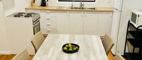 Ett modernt kök med vita skåp, en fläktkåpa i rostfritt stål, en inbyggd ugn, en diskho och en mikrovågsugn. Det finns ett matbord med stolar i rotting och en skål med frukt på bordet.