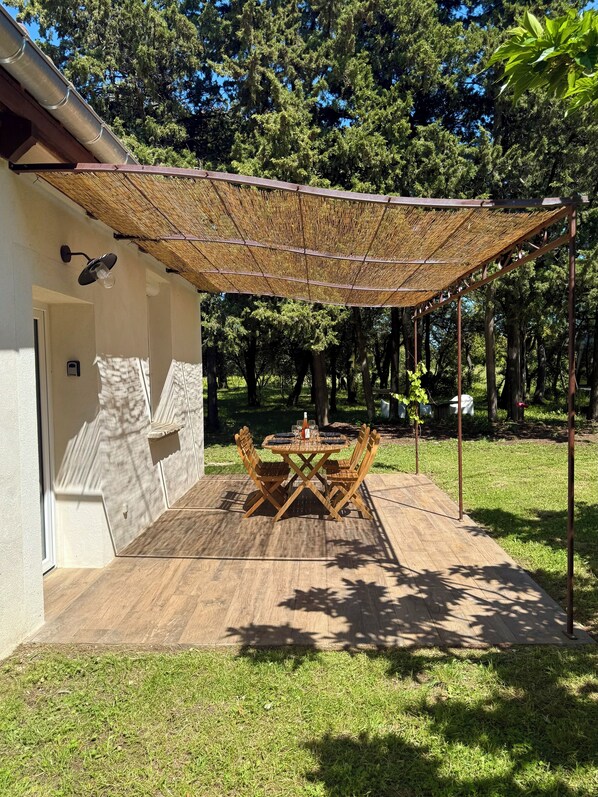 A covered patio with a table and chairs, surrounded by a grassy area and trees. - Maisonnette Avec Petite Forêt - Tranquillité Assurée - Idéal Jeune Famille (Valréas)