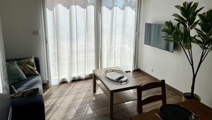 Una habitación con piso de madera, un sofá con cojines, una mesa de comedor con sillas, un televisor montado en la pared y una planta en maceta.