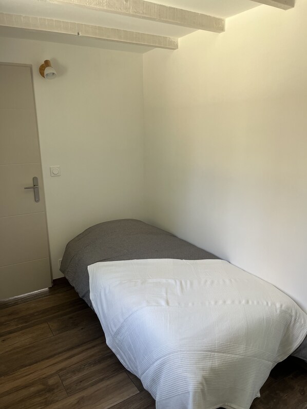 A small, single-bed room with a wooden floor, a white door, and a light switch on the wall. - Maisonnette Avec Petite Forêt - Tranquillité Assurée - Idéal Jeune Famille (Valréas)