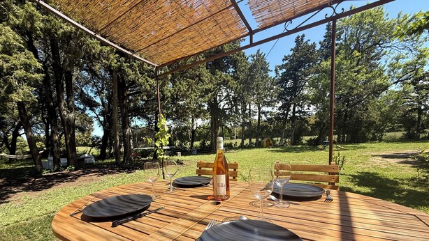 Mesa de madera al aire libre con platos, cubiertos y copas de vino, rodeada por un toldo de bambú.