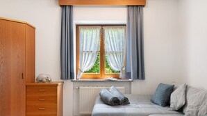 Une pièce avec une armoire en bois, une fenêtre avec des rideaux, un radiateur, un canapé recouvert de couvertures pliées et un bol posé sur le sol.
