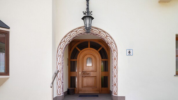 Une porte en bois surmontée d’un arc décoratif, une lampe en métal au-dessus, et une petite marche menant à l’entrée.