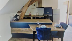 A dining area with a wooden table and blue chairs, a kitchen in the background, and exposed wooden beams on the ceiling.