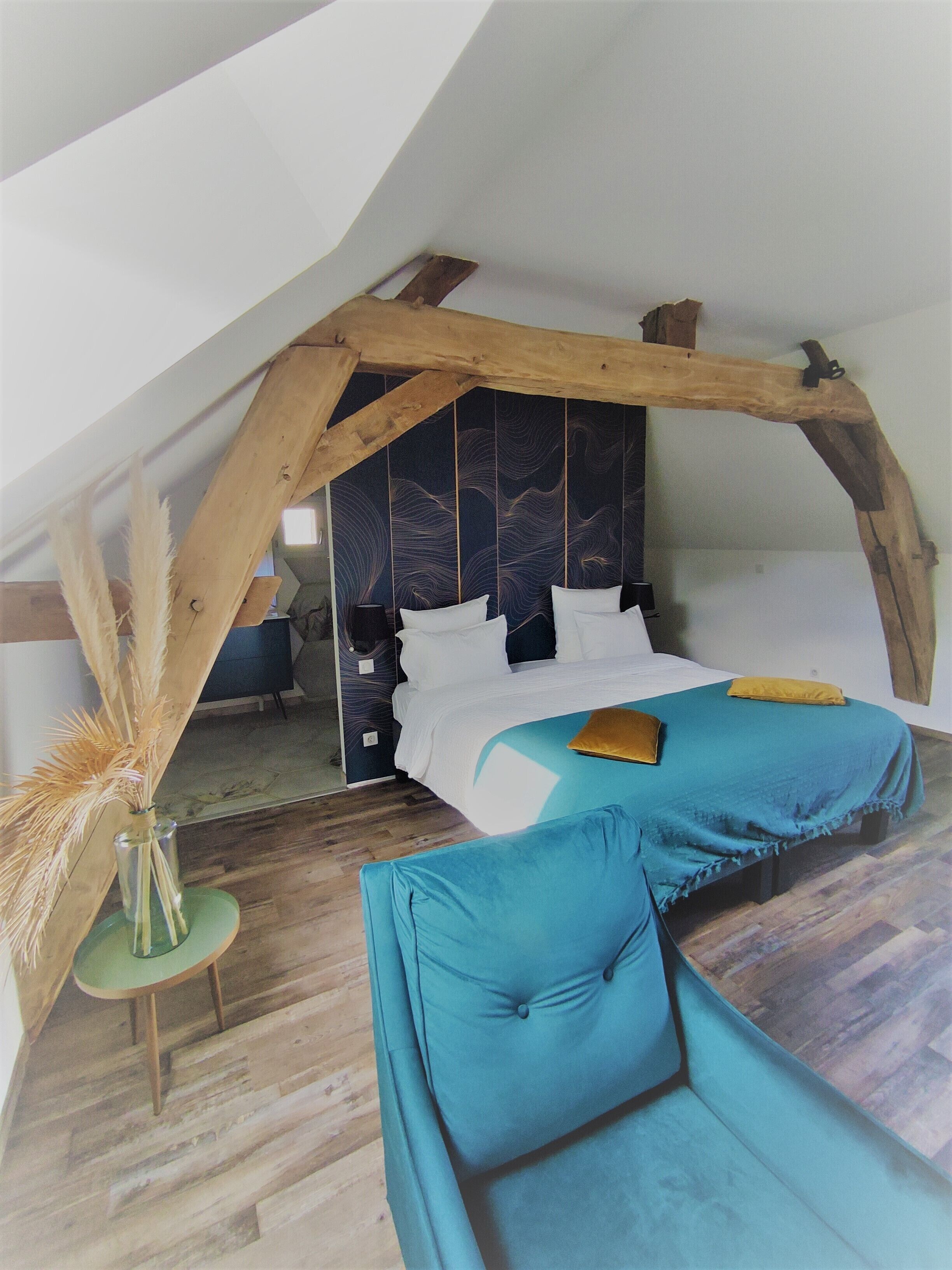A bedroom with a sloped ceiling, a bed with white bedding, a blue sofa, and a vase with dried plants.