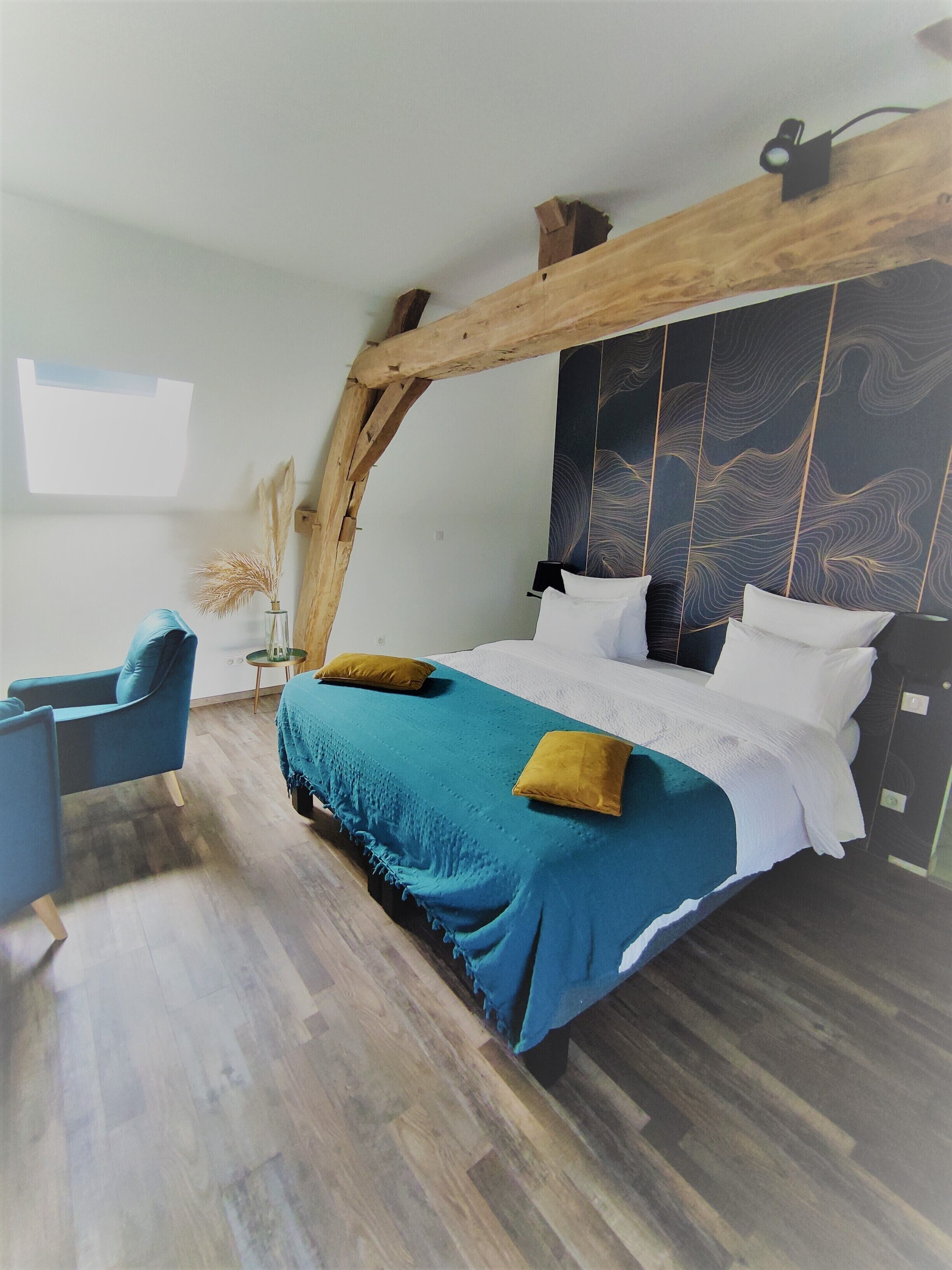 A modern bedroom with a wooden ceiling, a large bed with blue and white bedding, a blue armchair, and a small table with a vase of dried plants.