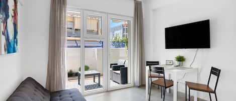 A modern living room with a sofa, a dining table with chairs, a TV mounted on the wall, and a sliding glass door leading to an outdoor area.