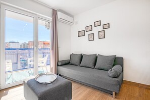 Un salon moderne avec un canapé gris, une petite table et un balcon offrant une vue sur les immeubles.