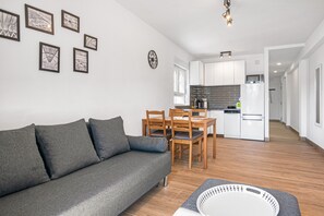 Un salon moderne avec un canapé gris, une table à manger et des chaises en bois, une cuisine avec des meubles blancs et une horloge au mur.