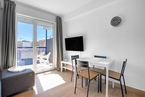 A modern room with a sofa, a dining table with chairs, a flat-screen TV, and a large window with curtains.
