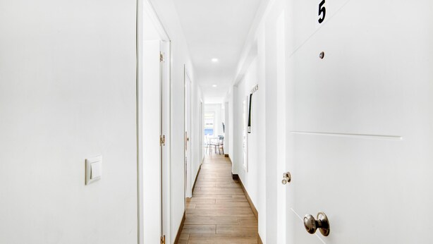 Un couloir aux murs blancs et au plancher en bois, qui mène à une pièce dont la porte est marquée du numéro 5.