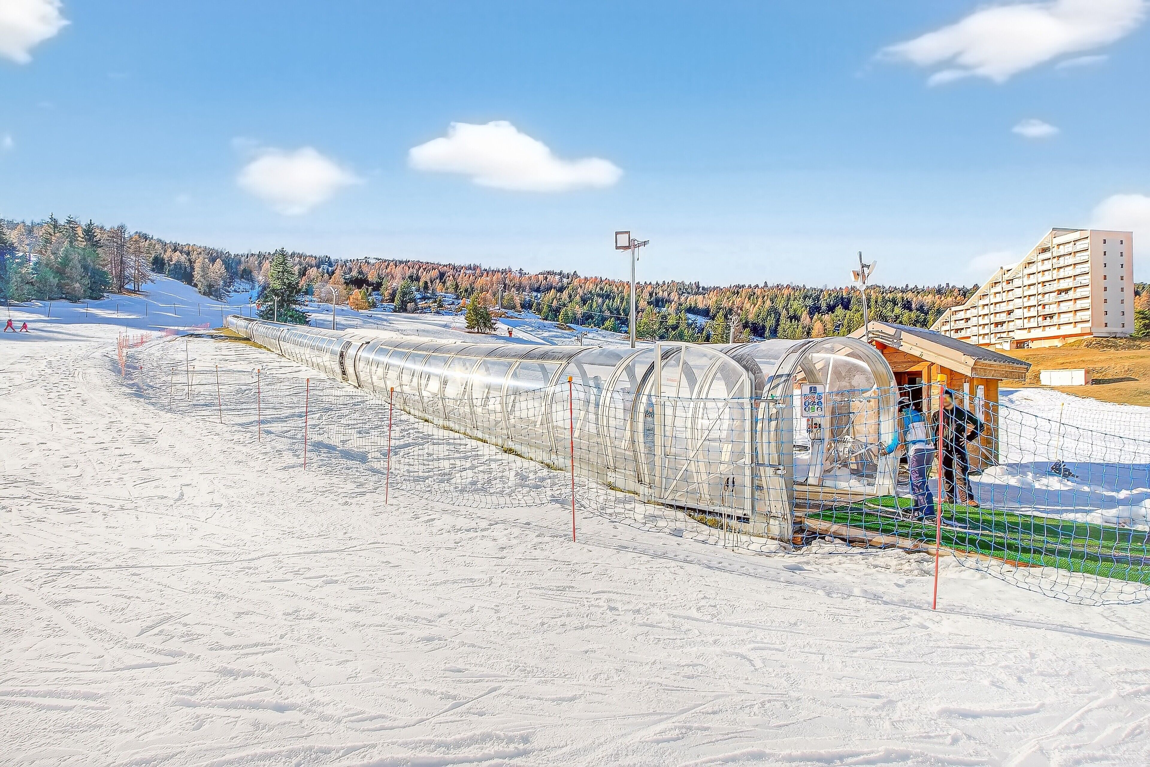 Une piste de ski enneigée, surmontée d’une structure transparente en forme de serre et d’un bâtiment doté d’un toit en pente.