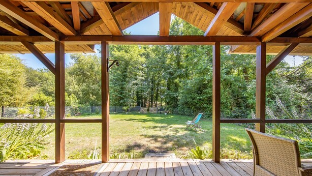 Une terrasse en bois avec un banc, une structure en forme de pergola et une vue sur une cour arrière verdoyante, agrémentée d’arbres.