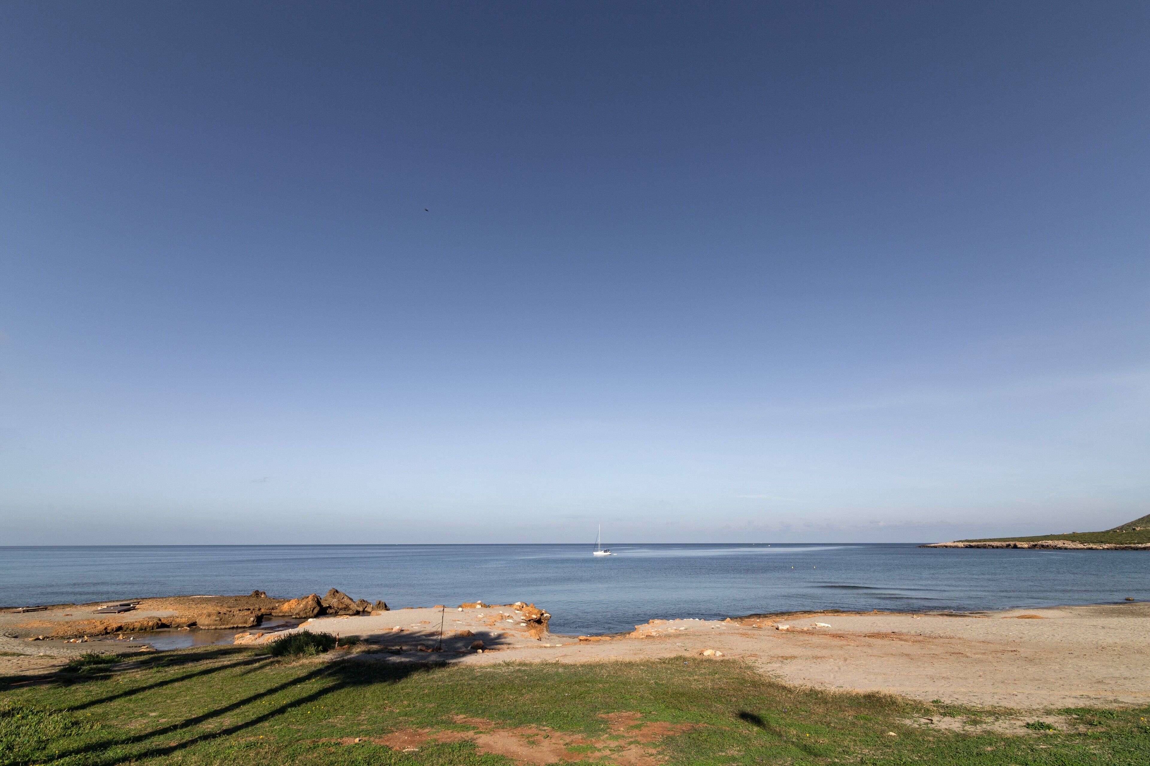 Paesaggio costiero con cielo sereno, spiaggia sabbiosa e una vela all'orizzonte.