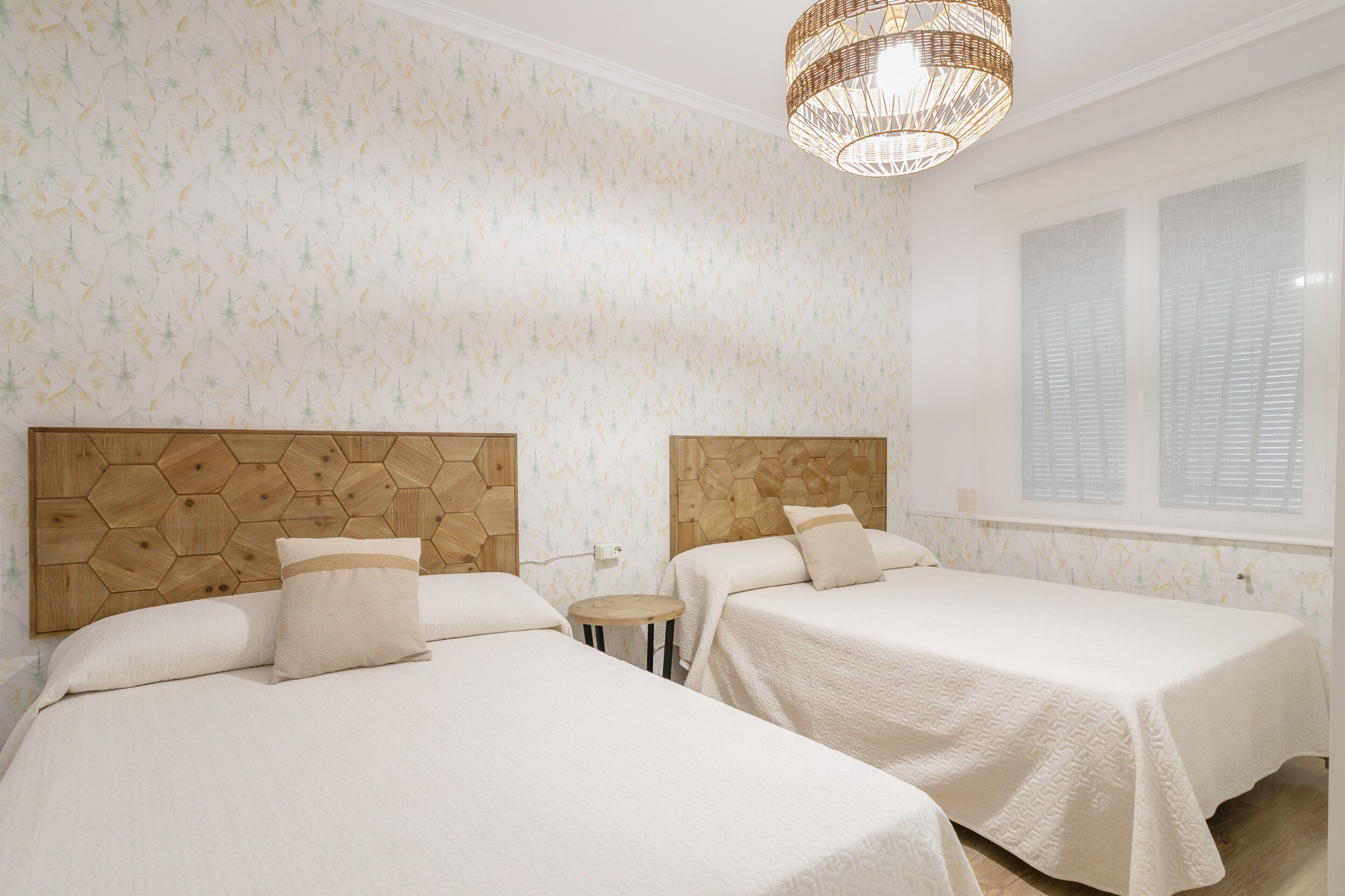 A hotel room with two beds, a wooden headboard, a small round table, and a large window with blinds.