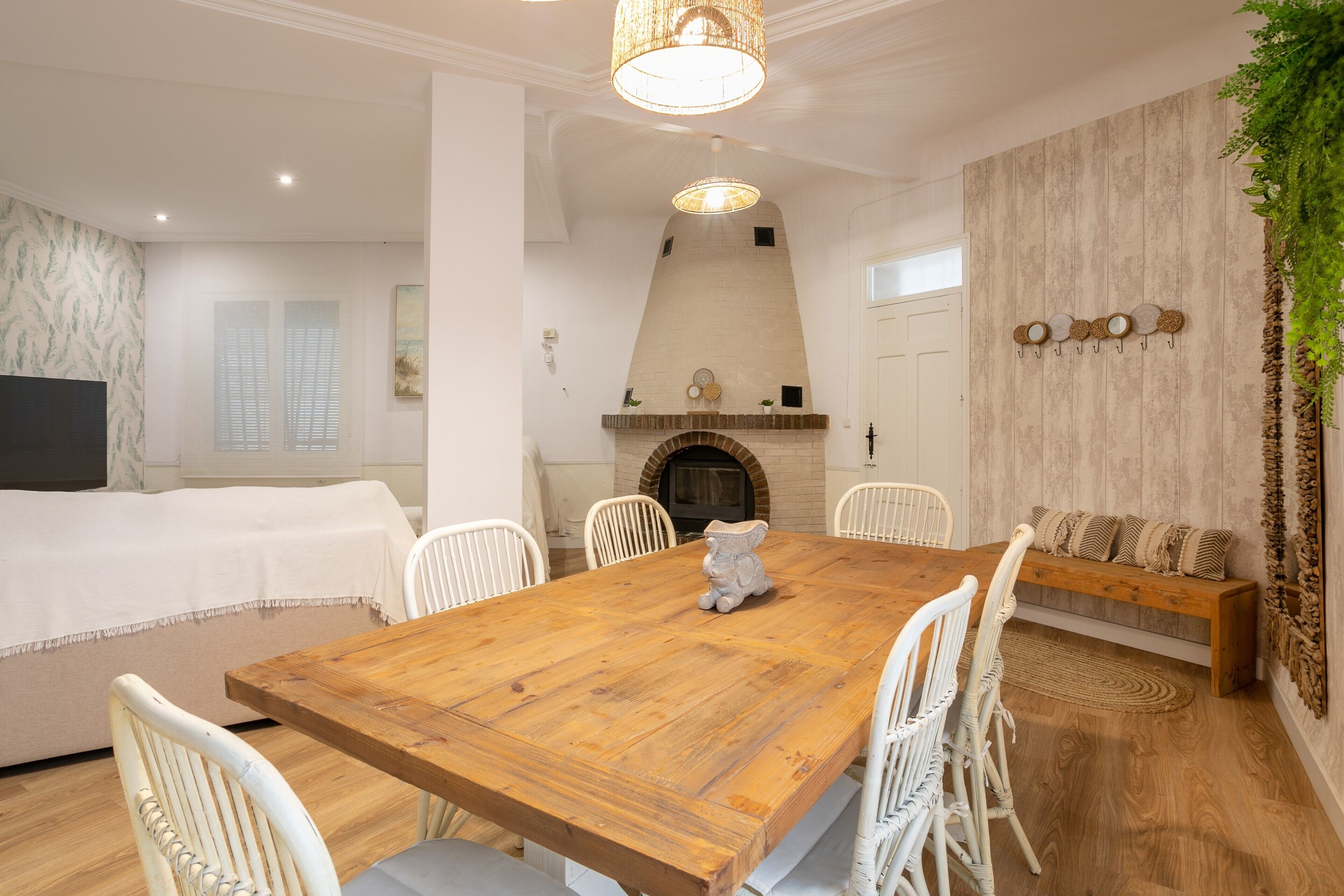 Un comedor con mesa de madera, sillas blancas, chimenea y una planta colgante.